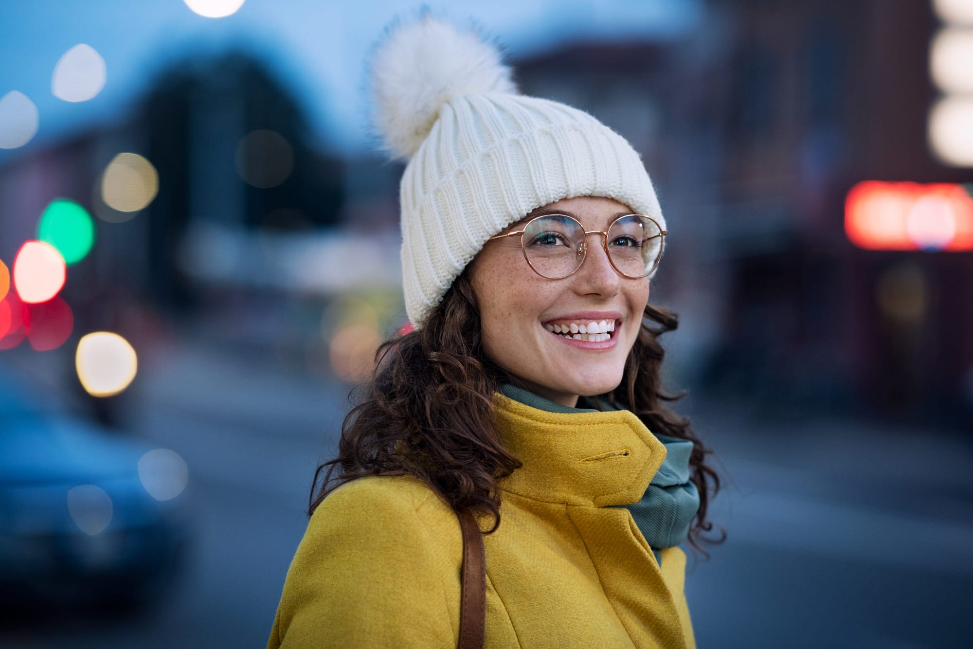 Smiling young woman wearing warm hat with eyeglasses on city street waiting for the bus.