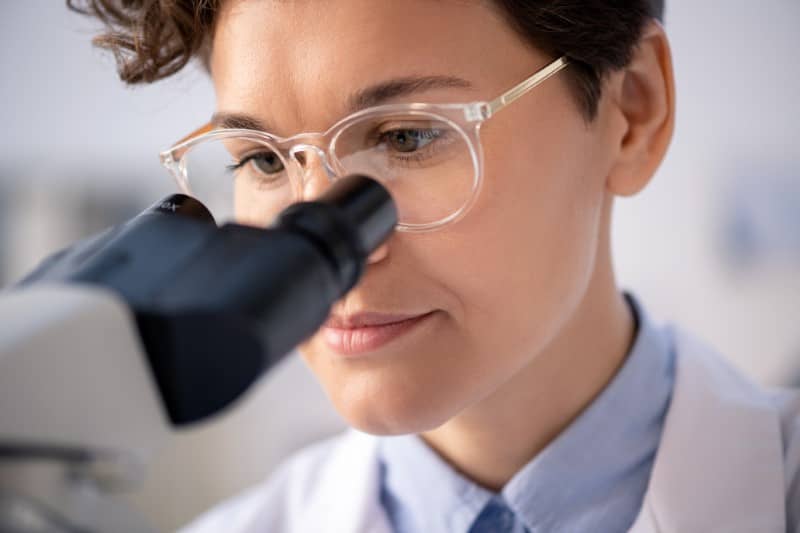 Head of young female scientist in eyeglasses and whitecoat studying chemical reaction of elements in microscope during research